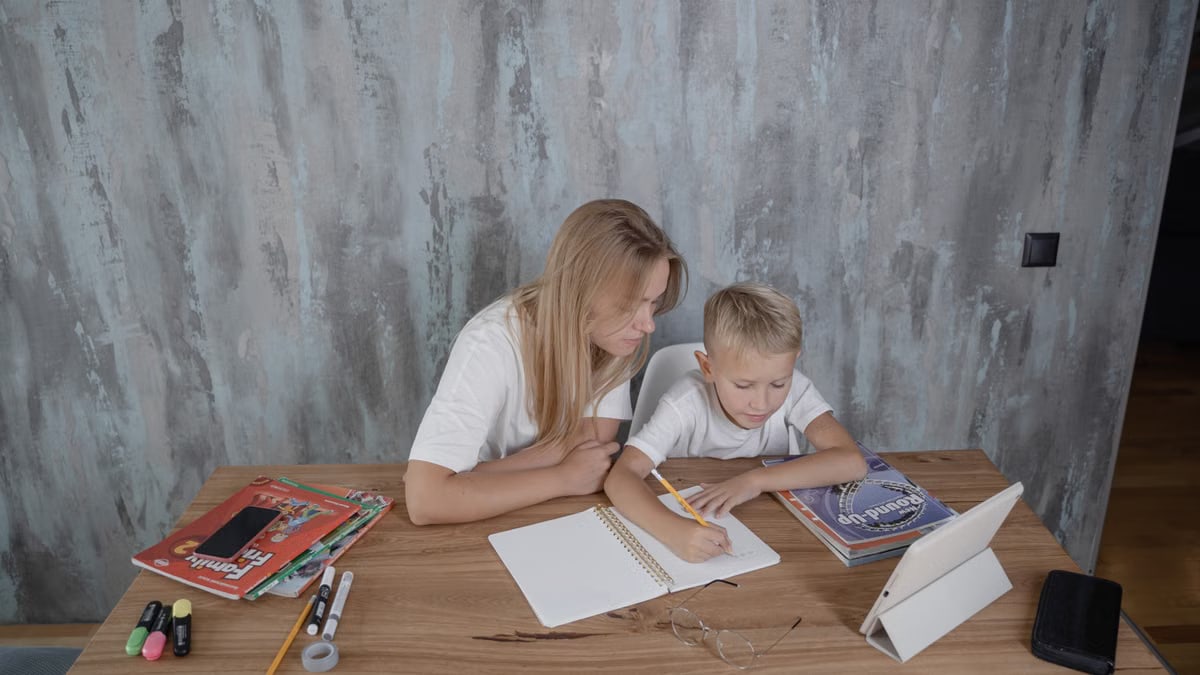 A mother guides her young son during a homework session at a desk in a cozy home setting.